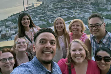 Un selfie de groupe de collègues souriants avec une vue de la ville en arrière-plan, pris depuis un point de vue élevé au coucher du soleil.